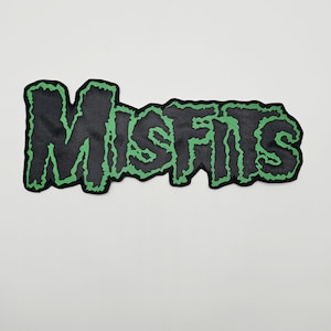 May include: A fabric patch featuring the word MISFITS in a stylized, jagged font. The lettering is black with a vibrant green outline, creating a high-contrast design. The edges of the patch follow the irregular, dripping shape of the text.