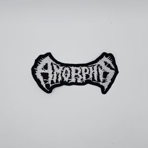 May include: A black and white embroidered patch with the word "AMORPHIS" in a stylized font. The patch has a unique shape with pointed edges and a black border. The text is in white.