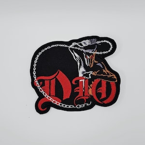 May include: Black embroidered patch with the word "DIO" in red gothic lettering. A white chain and a detailed illustration of a figure with orange and white accents are also present. The patch has a black border.