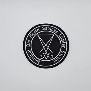May include: A black circular patch with white embroidered text and a central symbol. The text reads "Nomine Dei Nostri Satanas Lucifer Excelsi" around the edge. The central symbol is a stylized inverted pentagram.