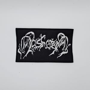 Puede incluir: Parche rectangular negro con texto blanco bordado "MOSKEEEN". El texto está en una fuente gótica estilizada. El parche es probablemente para ropa o accesorios.