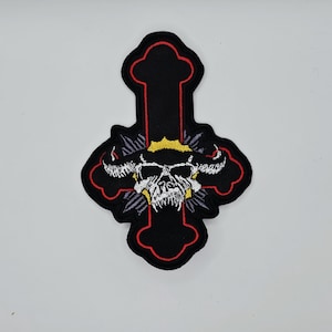 Op de afbeelding: Zwart geborduurde patch in de vorm van een kruis met een schedelontwerp. Het kruis heeft een rode omtrek en de schedel is wit met gele en paarse accenten. De patch is waarschijnlijk voor kleding of accessoires.