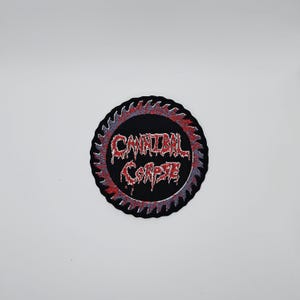 May include: A round, black patch with the text "CANNIBAL CORPSE" in red, dripping letters. The text is surrounded by a red and gray ring, and a black saw blade edge.