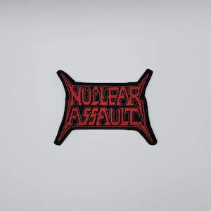 Puede incluir: Un parche negro con la inscripción "NUCLEAR ASSAULT" en rojo, con una tipografía estilizada. El parche tiene un borde angular y está diseñado para ser cosido o planchado en ropa.
