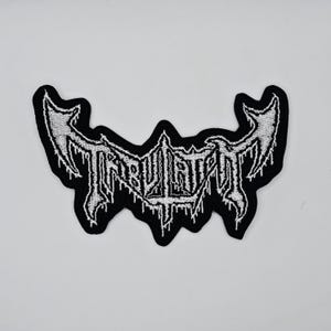 May include: A black and white embroidered patch with the word "TRIBULATE" in a gothic font. The patch has a unique shape with pointed edges and a dripping effect, suitable for jackets or bags.