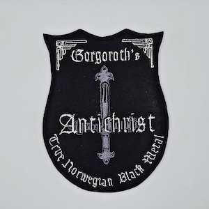 Może przedstawiać: Czarna naszywka z białym haftowanym tekstem i szarym krzyżem. Na naszywce widnieje napis "Gorgoroth's Antichrist True Norwegian Black Metal" gotycką czcionką. Naszywka ma kształt tarczy.