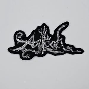 Puede incluir: Parche bordado negro con el texto "Atheist Blood" en fuente blanca estilizada. El parche tiene una forma irregular y está diseñado para ser cosido en ropa o accesorios.