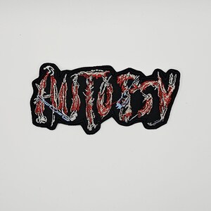 May include: An embroidered patch featuring the word AUTOPSY in a stylized, jagged font. The lettering is rendered in shades of red, white, and light blue, designed to resemble raw muscle tissue and bone, set against a solid black background.