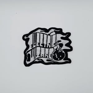 Peut inclure: Écusson brodé noir et blanc avec le texte "ELECTRIC WIZARD" dans une police stylisée. Le patch présente une bordure noire avec un motif ondulé et un graphique de bombe.