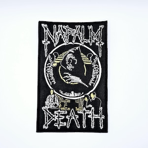 May include: Black rectangular patch with white embroidered text and graphics. The text "NAPALM DEATH" is at the top, with a circular graphic of a skull and skeleton. The word "DEATH" is at the bottom, with two figures on either side.