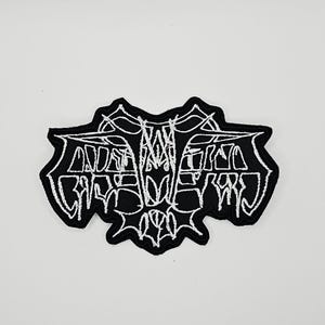 May include: A black embroidered patch featuring a white, stylized, symmetrical design with sharp, angular lines and intricate, web-like patterns. The embroidery is dense, creating a high-contrast graphic element with a distinct, jagged border shape.