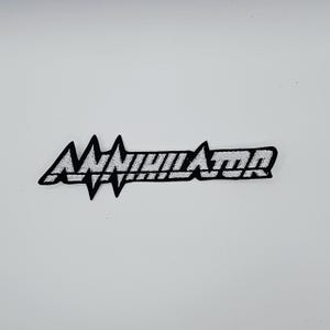 May include: A black and white embroidered patch with the word "ANNIHILATOR" in a stylized font. The patch has a jagged design element on the left side and is likely for clothing or accessories.