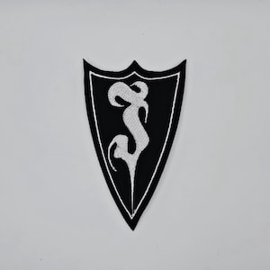 Peut inclure: Écusson brodé noir et blanc en forme de bouclier. L'écusson présente un « S » blanc stylisé sur fond noir, avec une bordure blanche. L'écusson est probablement destiné aux vêtements ou aux accessoires.