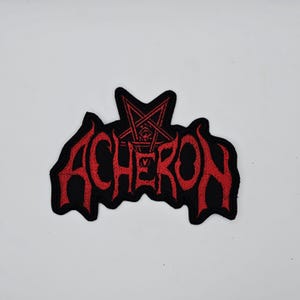 Może przedstawiać: Czarna, haftowana naszywka z napisem "ACHERON" w czerwonych, stylizowanych gotyckich literach. Czerwony pentagram znajduje się nad słowem. Naszywka ma kształt nietoperza.