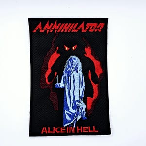 May include: Black rectangular patch with the word "ANNIHILATOR" in red, above a blue and white illustration of a figure holding a doll and candle. The text "ALICE IN HELL" is below the illustration. A red silhouette of a demon is behind the figure.