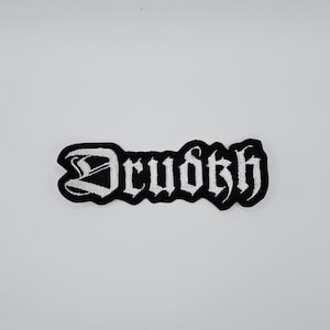 May include: A black and white embroidered patch featuring the word "Drudkh" in a gothic font. The patch has a black border and the text is in white. The patch is rectangular and is designed to be sewn or ironed onto clothing or accessories.