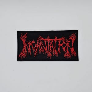 May include: A rectangular black patch with red embroidered text. The text appears to be a band name in a gothic font. The patch is likely for a jacket or bag.