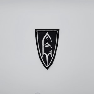 May include: A black and white embroidered patch in a shield shape. The patch features a stylized design of a face with an eye and a gothic arch. The patch has a black background with a white border.