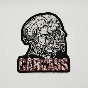 Op de afbeelding: Een geborduurde patch met een zwart-wit anatomisch profiel van een menselijk hoofd met blootliggend hersen- en spierweefsel. Onder het hoofd staat het woord CARCASS geborduurd in witte blokletters met een dunne rode omlijning op een zwarte achtergrond.