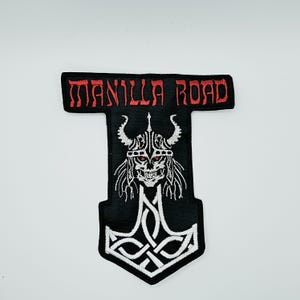 May include: A black embroidered patch shaped like a hammer, featuring the red text MANILLA ROAD at the top. Below the text is a white embroidered graphic of a horned skull with red eyes, set above an intricate white Celtic knotwork design.