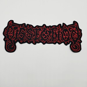 Op de afbeelding: Een rechthoekige geborduurde patch met het woord DISSECTION in een gestileerd, grillig, vlamachtig lettertype. Het ontwerp bestaat uit zwarte letters omlijnd met felrood garen, wat zorgt voor een contrastrijke en scherpe grafische uitstraling.