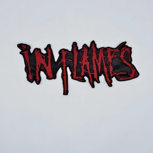 May include: A black patch with the word "IN FLAMES" in red, stylized lettering. The patch has a jagged, distressed edge, and the text appears to be dripping or melting. The background is plain white.