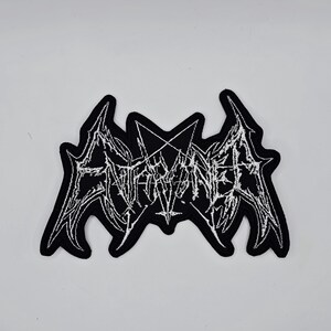 May include: Black patch with white embroidered text reading "ENTHRONED" in a gothic font. The patch has a unique shape with pointed edges and a pentagram design.