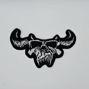 Może przedstawiać: Czarna i biała naszywka z haftem przedstawiająca czaszkę z rogami. Czaszka jest szczegółowo wyhaftowana białymi nićmi, podkreślając zęby i oczodoły, na czarnym tle. Naszywka ma charakterystyczny kształt z zakrzywionymi rogami.