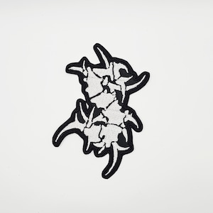 Puede incluir: Un parche bordado en blanco y negro con un diseño abstracto. El parche tiene un borde negro y un interior blanco texturizado. El diseño es una forma estilizada y simétrica con ángulos y curvas.