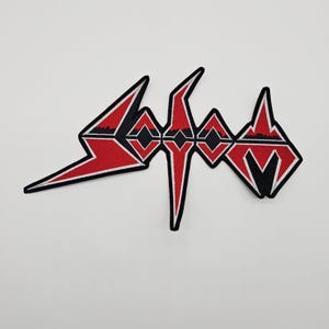 May include: Embroidered patch with the word "Sodom" in a distinctive, angular font. The patch is red and black with white edging. The design is striking and graphic, ideal for application on jackets, bags, or other fabric items.