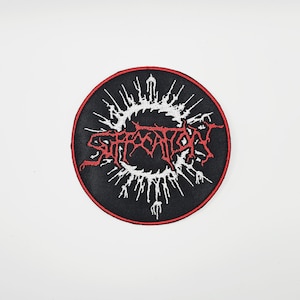 Può includere: Toppa rotonda nera con bordo rosso. La toppa presenta la scritta "SUFFOCATION" in rosso, con un lettering stilizzato, circondata da un disegno bianco a punte. La toppa è probabilmente per merchandising di band o musica.