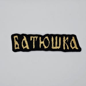 May include: Black embroidered patch with the word "БАТЮШКА" in gold lettering. The patch has a rectangular shape with a black border and is likely designed to be sewn onto clothing or accessories.