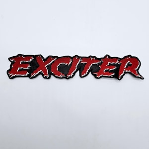 May include: A black patch featuring the word "EXCITER" in red, distressed lettering. The patch has a jagged edge, and the text is slightly raised from the black background. The overall design has a bold, edgy aesthetic.