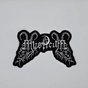 以下が含まれることがあります： ゴシック体で「Mysticuff」の文字が刺繍された白黒のパッチ。パッチは、雄羊の角と三叉のシンボルをあしらった様式化されたデザインが特徴です。パッチの幅は約10cm、高さは約5cmです。