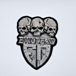 Puede incluir: Parche bordado con tres calaveras sobre un escudo. El parche tiene el texto "EVILDEAD" y un diseño estilizado. El parche es blanco y negro.