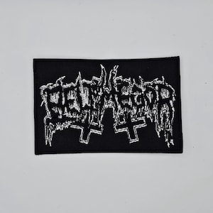 Puede incluir: Parche rectangular negro con texto bordado en blanco y dos cruces invertidas. El texto parece ser el nombre o logotipo de una banda, con una estética gótica o metal. El parche es para ropa o accesorios.