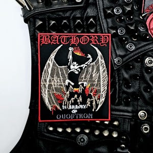 Op de afbeelding: Zwarte patch met rode rand met het woord "BATHORY" boven een gevleugelde figuur met vlammen en de tekst "IN MEMORY OF QUORTHON". De patch is genaaid op een zwart leren jasje met zilveren studs en spikes.