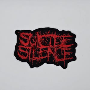 Könnte beinhalten: Schwarzer, gestickter Aufnäher mit den Worten "SUICIDE SILENCE" in roter, stilisierter, gotischer Schrift. Der Aufnäher hat einen gezackten, unregelmäßigen Rand. Der Text ist im Stil einer Heavy-Metal-Band gehalten.