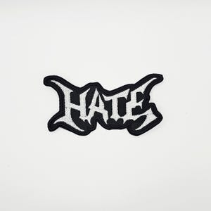May include: A black and white embroidered patch with the word "HATE" in a gothic font. The patch has a black border and is set against a white background. The letters are white.
