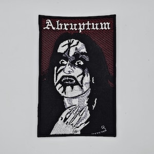 May include: Embroidered patch featuring a black and white image of a person with dramatic face paint. The patch has the word "Abruptum" in a gothic font at the top. The background is a dark red.