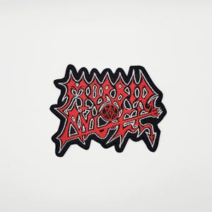 May include: Embroidered patch with a red and black design. The patch features a stylized, jagged outline with a central design in red, white, and black. The design includes a pentagram and abstract shapes, suitable for jackets or bags.