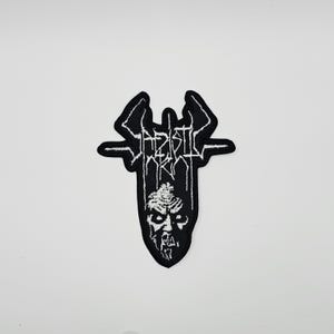 May include: Black and white embroidered patch featuring the text "Baptist Rot" in a stylized font, with a demonic face at the bottom. The patch has a unique, symmetrical shape with wing-like elements.