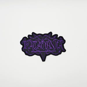 May include: A black patch with the word "HADACONIA" embroidered in purple, featuring a stylized, gothic font. The patch has a unique, irregular shape with pointed edges and a decorative border, suitable for clothing or accessories.