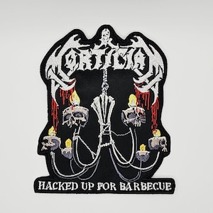 May include: A black embroidered patch featuring the white text Mortician at the top and Hacked Up For Barbecue at the bottom. The center design depicts a silver chandelier with skulls and lit candles dripping with red blood-like accents.