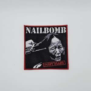 Könnte beinhalten: Quadratischer schwarzer Aufnäher mit rotem Rand. Der Aufnäher zeigt das Wort "NAILBOMB" über einer Schwarz-Weiß-Grafik einer Person. Der Text "POINT BLANK" befindet sich unter der Grafik.