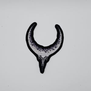 以下が含まれることがあります： 黒い縁取りのある三日月デザインの刺繍パッチ。 月は、テクスチャ加工された薄い灰色のパターンと、下部に滴り落ちる効果で満たされています。 パッチは白い背景にあります。