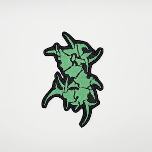 May include: A green and black embroidered patch featuring an abstract design. The patch has a jagged, organic shape with a black border and a green interior. The patch is likely for clothing or accessories.