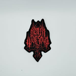 Puede incluir: Un parche bordado negro con el texto rojo "DEATH WORSHIP" en una fuente estilizada y dentada. El diseño incluye una forma central similar a una cruz con un motivo de calavera en la base, todo delineado con hilo negro sobre fondo negro.