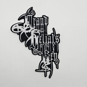 Puede incluir: Parche bordado en blanco y negro con el texto "Grand Rituals". El parche presenta un diseño de fuente gótica estilizada. El parche está sobre un fondo blanco.