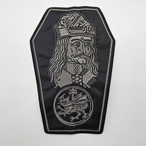 Può includere: Una toppa ricamata nera e grigia a forma di bara. La toppa presenta un ritratto di un uomo con una corona e un drago con le ali.